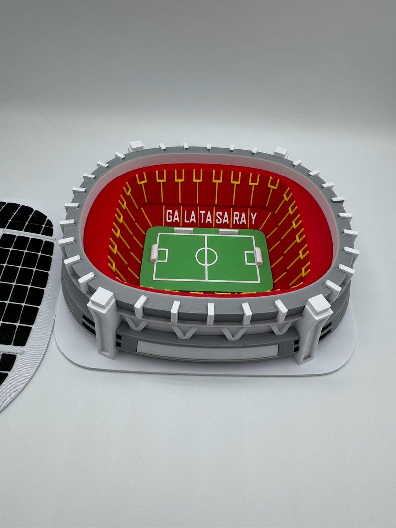 May include: A miniature stadium model with a green field and the text "GALA TA SARAY" in the center. The stadium has red seating, gray walls, and white accents. The model is a detailed representation of a soccer stadium.