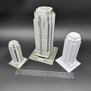 May include: Three white and gray miniature skyscraper models of varying sizes. The tallest model is centered, flanked by two smaller models. A ruler is placed in front of the models, providing scale. The buildings have detailed architectural features.