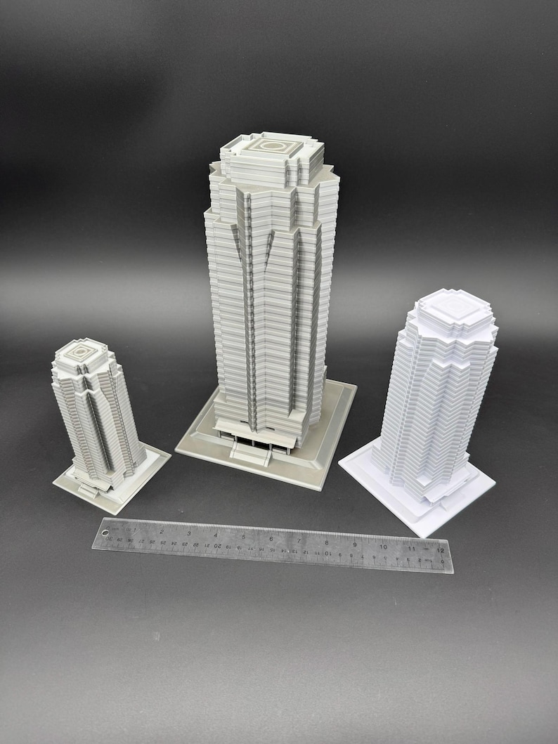 May include: Three white and gray miniature skyscraper models of varying sizes. The tallest model is centered, flanked by two smaller models. A ruler is placed in front of the models, providing scale. The buildings have detailed architectural features.