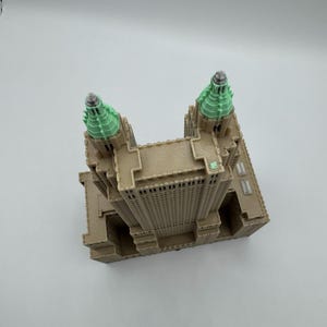 Może przedstawiać: Szczeg&oacute;łowy model miniaturowy wieżowca, z fasadą w kolorze beżowym i wieżami zwieńczonymi zielenią. Model prezentuje detale architektoniczne, w tym okna i elementy dekoracyjne, na białym tle.