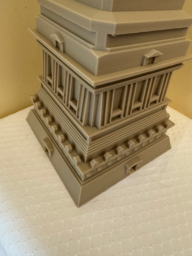 May include: A 3D printed model of a multi-level structure with a square base and a pointed top. The model is made of a light brown plastic and has intricate details, including columns, arches, and a decorative border.