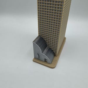 May include: A 3D-printed model of a skyscraper with a tan facade and a gray base. A smaller gray structure is attached to the base of the building. The model is set against a white background.