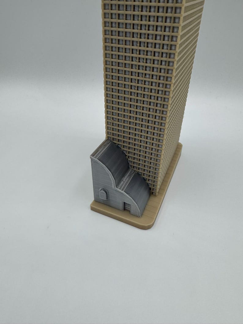 May include: A 3D-printed model of a skyscraper with a tan facade and a gray base. A smaller gray structure is attached to the base of the building. The model is set against a white background.