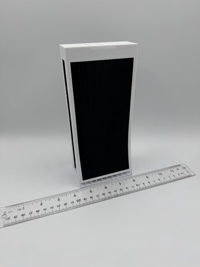 May include: A white 3D printed model of a tall building with a black facade. The model is approximately 7 inches tall and is sitting on a white surface next to a ruler.