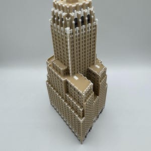 May include: A detailed model of a skyscraper, with a tan exterior and white detailing. The building has a tiered design, with a rectangular base and a tall, slender tower. The model is set against a plain white background.