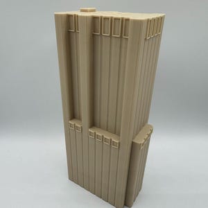 Puede incluir: Maqueta arquitectónica beige de un rascacielos. El edificio tiene líneas verticales y detalles rectangulares, con un techo plano y una sección más pequeña en la base. El modelo está hecho de un material sólido y se alza verticalmente.