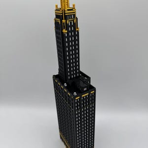 Op de afbeelding: Een zwart en goudkleurig wolkenkrabbermodel met een gedetailleerd ontwerp. Het gebouw heeft een rechthoekige basis en een hoge, slanke toren. De bovenkant van de toren is goudkleurig en de ramen zijn wit. Het model staat tegen een witte achtergrond.