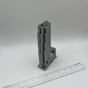 May include: A gray 3D printed model of a skyscraper building. The model is about 7 inches tall and is sitting on a white surface. A ruler is in the foreground.