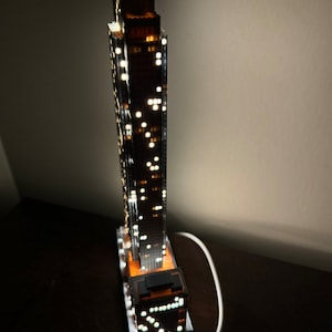 May include: A detailed model of a skyscraper with illuminated windows, lit from within, and a white cord plugged into an outlet.