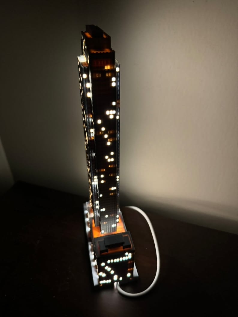 May include: A detailed model of a skyscraper with illuminated windows, lit from within, and a white cord plugged into an outlet.