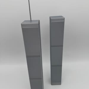 Può includere: Un modello in scala stampato in 3D delle Torri Gemelle di colore grigio. La torre più alta a sinistra presenta una sottile antenna che si estende dal tetto. Entrambe le strutture rettangolari mostrano un motivo a griglia sulla facciata.