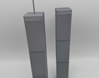 May include: A gray 3D printed scale model of the Twin Towers. The taller tower on the left features a thin antenna spire extending from the roof. Both rectangular structures display a repeating grid pattern representing the exterior facade.