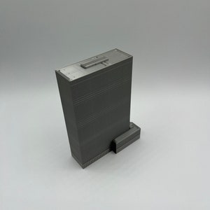 May include: A gray 3D printed model of a skyscraper with a flat roof and a small rectangular building attached to the side.