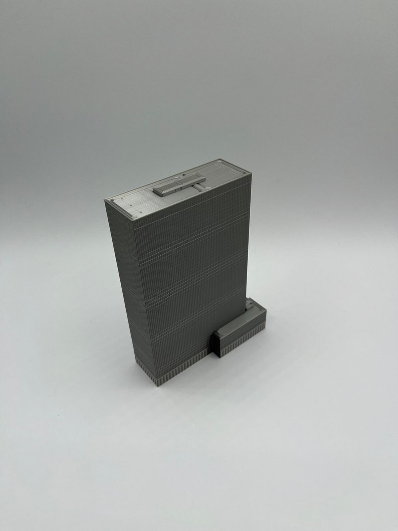 May include: A gray 3D printed model of a skyscraper with a flat roof and a small rectangular building attached to the side.