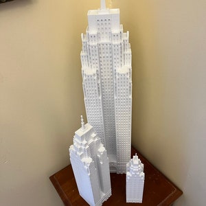 May include: Three white 3D printed models of skyscrapers, the tallest is about 10 inches tall, the middle is about 6 inches tall, and the smallest is about 4 inches tall. The models are on a wooden surface.