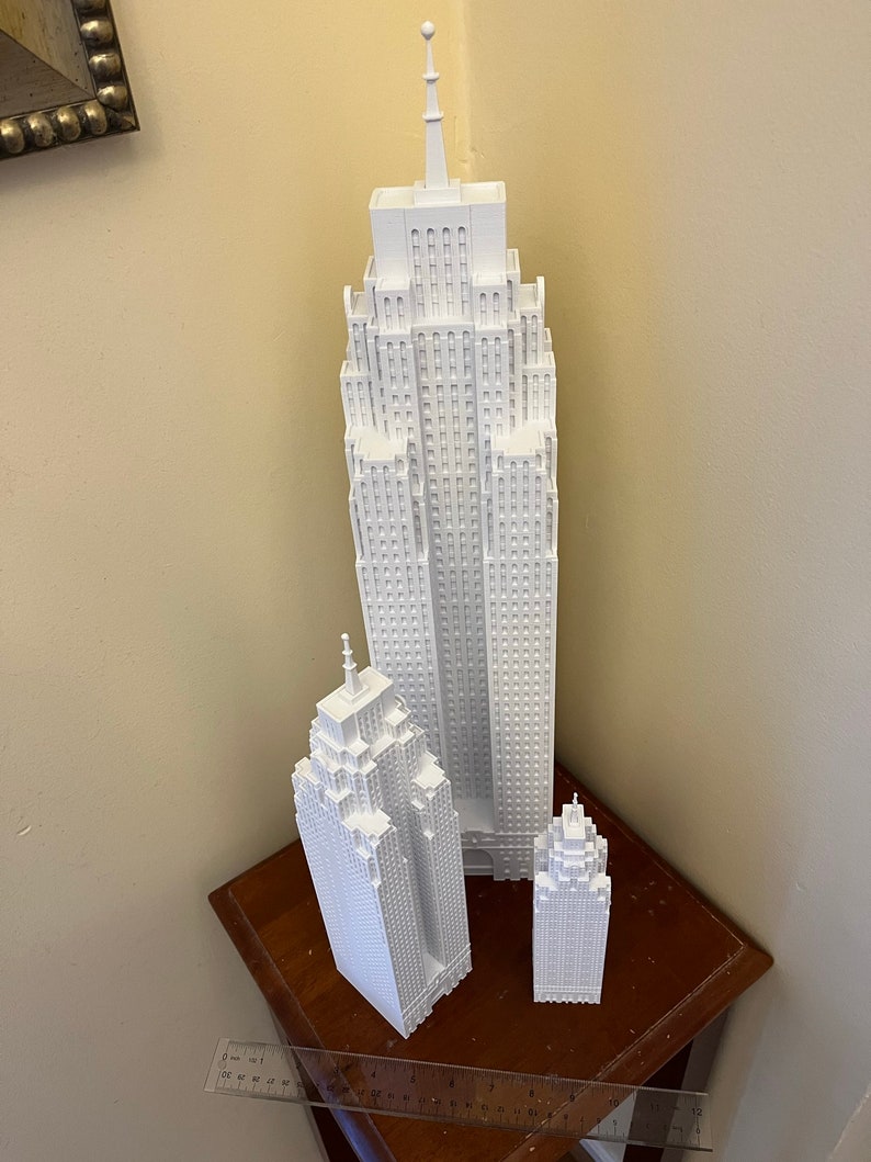 May include: Three white 3D printed models of skyscrapers, the tallest is about 10 inches tall, the middle is about 6 inches tall, and the smallest is about 4 inches tall. The models are on a wooden surface.