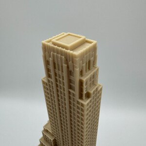 May include: A beige, miniature skyscraper model with a detailed facade. The building has a stepped design and a flat roof. The model is made of a solid material and is likely a decorative item or architectural model.