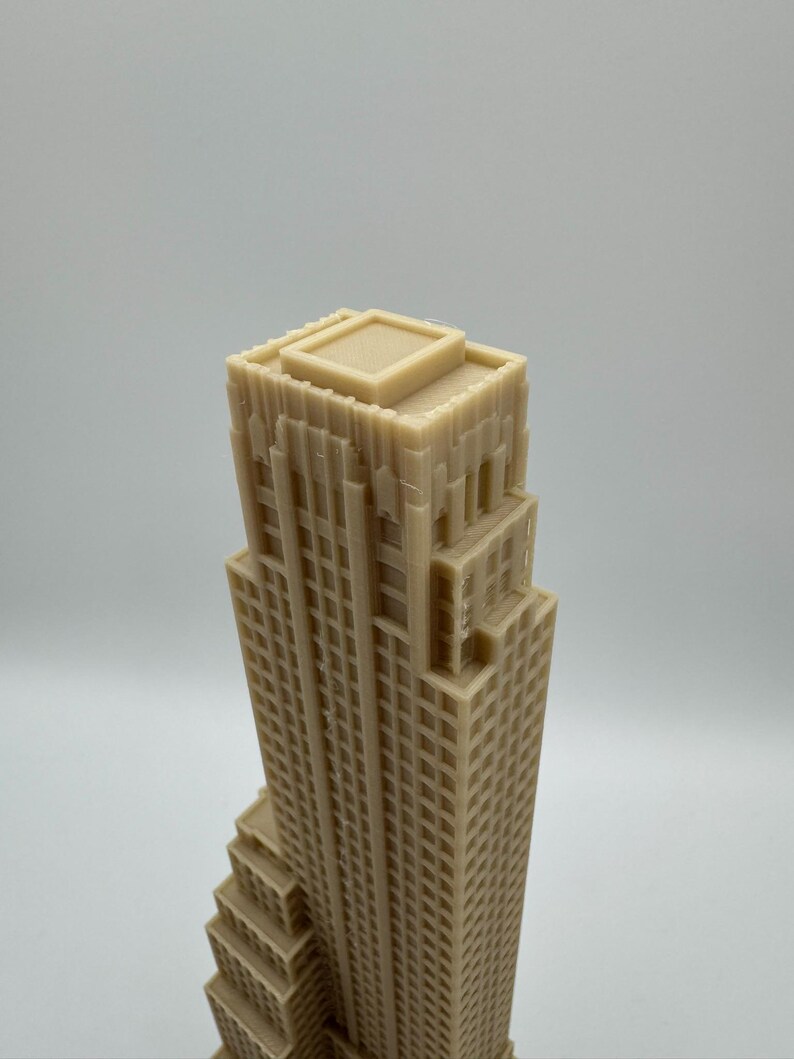 May include: A beige, miniature skyscraper model with a detailed facade. The building has a stepped design and a flat roof. The model is made of a solid material and is likely a decorative item or architectural model.