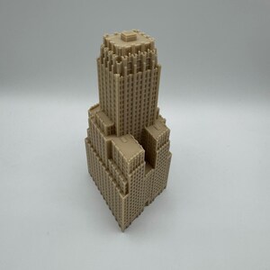 May include: A tan-colored architectural model of a skyscraper. The building has a tiered design with multiple setbacks and a flat roof. The model is detailed with numerous small windows and architectural features, showcasing a classic skyscraper design.