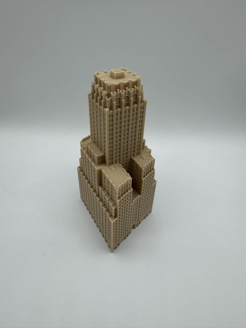 May include: A tan-colored architectural model of a skyscraper. The building has a tiered design with multiple setbacks and a flat roof. The model is detailed with numerous small windows and architectural features, showcasing a classic skyscraper design.
