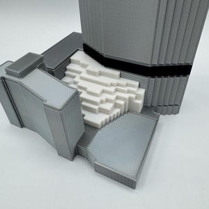 May include: A 3D-printed architectural model in shades of gray and white. The model features a tall, rectangular structure with vertical lines, a black band, and a textured base. The design includes a stepped white section.