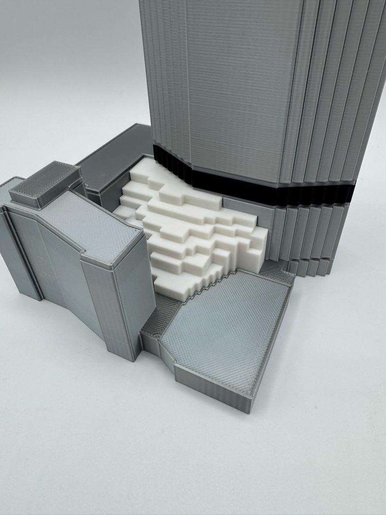 May include: A 3D-printed architectural model in shades of gray and white. The model features a tall, rectangular structure with vertical lines, a black band, and a textured base. The design includes a stepped white section.