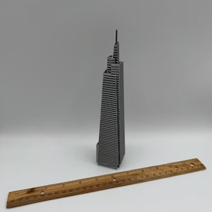 May include: A gray miniature skyscraper model with a ruler. The building has a detailed facade with many windows. The wooden ruler is marked with inch measurements from 1 to 30.