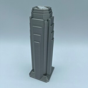 May include: A 3D printed model of a tall, gray skyscraper with a stepped design. The model is made of a smooth, gray plastic and has a detailed facade.