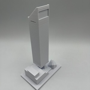 May include: A white 3D printed model of a skyscraper with a smaller building in front of it.