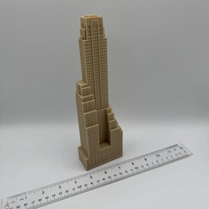 May include: A tan-colored miniature skyscraper model with a detailed facade. A clear ruler with inch markings is placed next to the building. The model appears to be approximately 10 inches tall.