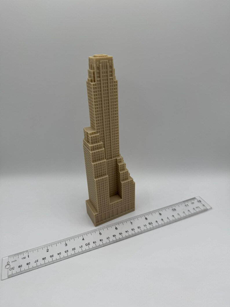 May include: A tan-colored miniature skyscraper model with a detailed facade. A clear ruler with inch markings is placed next to the building. The model appears to be approximately 10 inches tall.