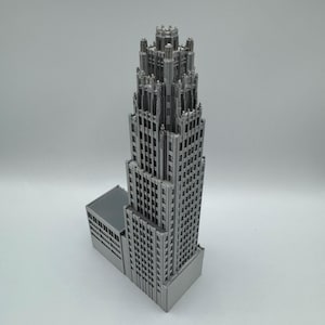 May include: A detailed, silver-colored miniature skyscraper model. The building features a tiered design with intricate architectural details, including numerous windows and a decorative top. The model is set against a plain white background, highlighting its structure.
