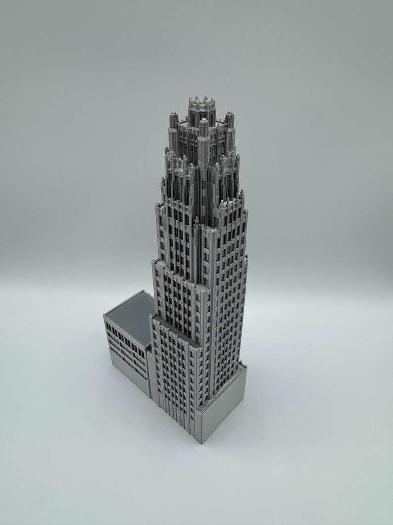 May include: A detailed, silver-colored miniature skyscraper model. The building features a tiered design with intricate architectural details, including numerous windows and a decorative top. The model is set against a plain white background, highlighting its structure.