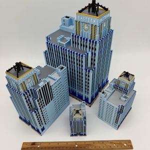 May include: A collection of light blue miniature buildings with the text "EASTERN" on the top. The buildings have black and gold accents. A wooden ruler is placed at the bottom of the image.