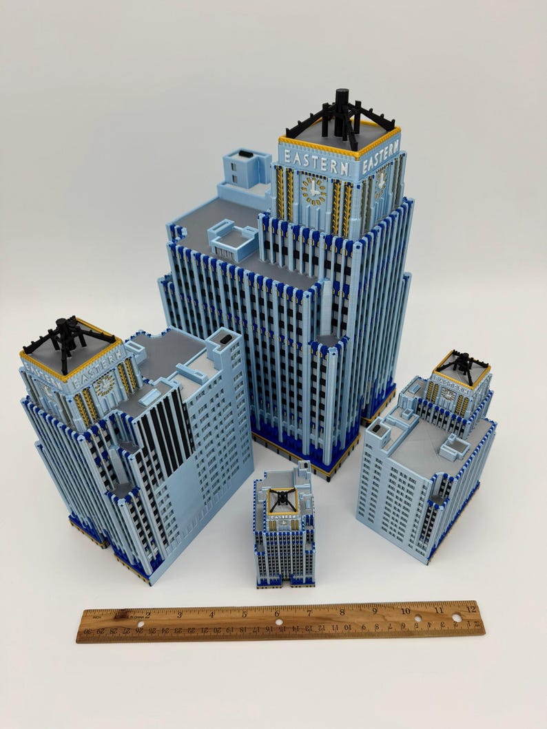 May include: A collection of light blue miniature buildings with the text "EASTERN" on the top. The buildings have black and gold accents. A wooden ruler is placed at the bottom of the image.