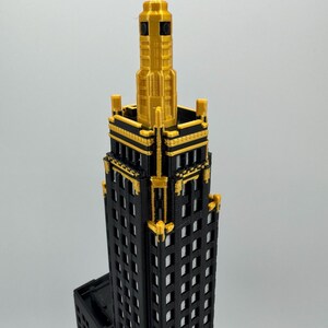 Op de afbeelding: Een 3D-geprint model van een wolkenkrabber. Het gebouw is zwart met gouden details, inclusief het bovenste gedeelte. De ramen zijn rechthoekig en gelijkmatig verdeeld. Het model staat tegen een effen achtergrond.