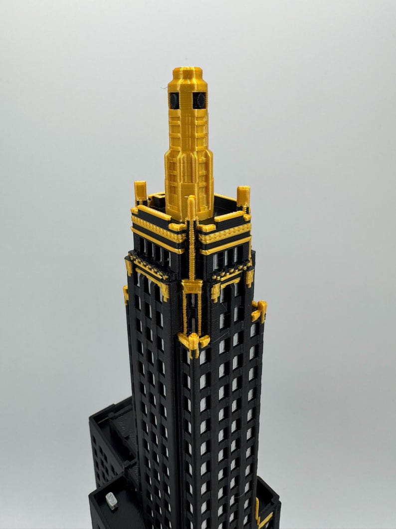 Op de afbeelding: Een 3D-geprint model van een wolkenkrabber. Het gebouw is zwart met gouden details, inclusief het bovenste gedeelte. De ramen zijn rechthoekig en gelijkmatig verdeeld. Het model staat tegen een effen achtergrond.