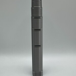 May include: A gray, miniature model of a tall skyscraper. The building has a rectangular base and a slender, multi-story design with numerous windows and horizontal lines. The top of the building has a detailed, textured roof.