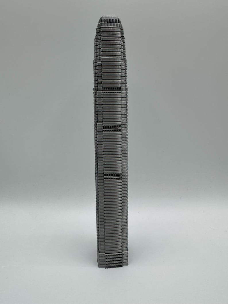 May include: A gray, miniature model of a tall skyscraper. The building has a rectangular base and a slender, multi-story design with numerous windows and horizontal lines. The top of the building has a detailed, textured roof.
