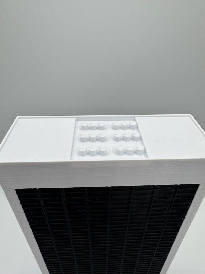 May include: White building model with a black grid pattern on the front and a white roof with a pattern of small white circles.