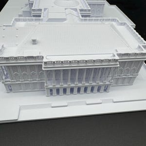 K&ouml;nnte beinhalten: Ein detailliertes, wei&szlig;es 3D-gedrucktes Modell eines Geb&auml;udes mit Kuppel, S&auml;ulen und mehreren Ebenen. Das Modell zeigt architektonische Merkmale, darunter ein Kuppeldach, Balkone und aufwendige Details. Das Modell ist vor einem dunklen Hintergrund platziert.