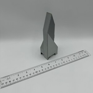 May include: A gray 3D printed model of a building with a pointed roof. The model is about 5 inches tall and is sitting on a white surface next to a ruler.