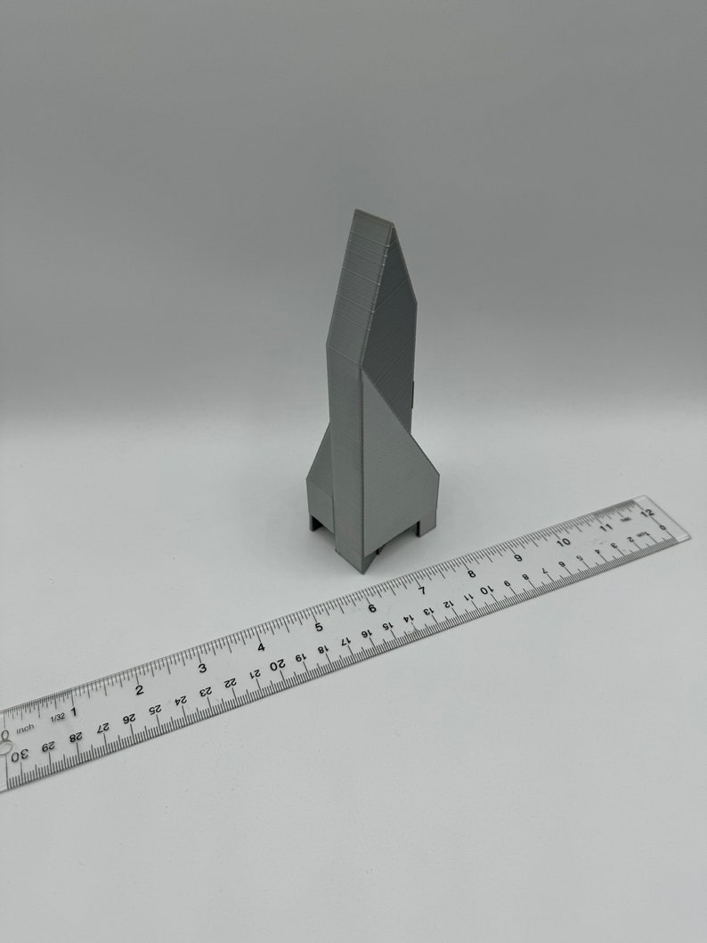 May include: A gray 3D printed model of a building with a pointed roof. The model is about 5 inches tall and is sitting on a white surface next to a ruler.