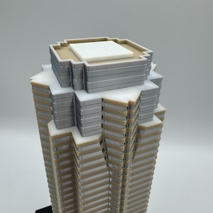 May include: A detailed architectural model of a skyscraper. The model features a layered design with alternating beige and gray sections, topped with a white and gray roof structure. The base is black.