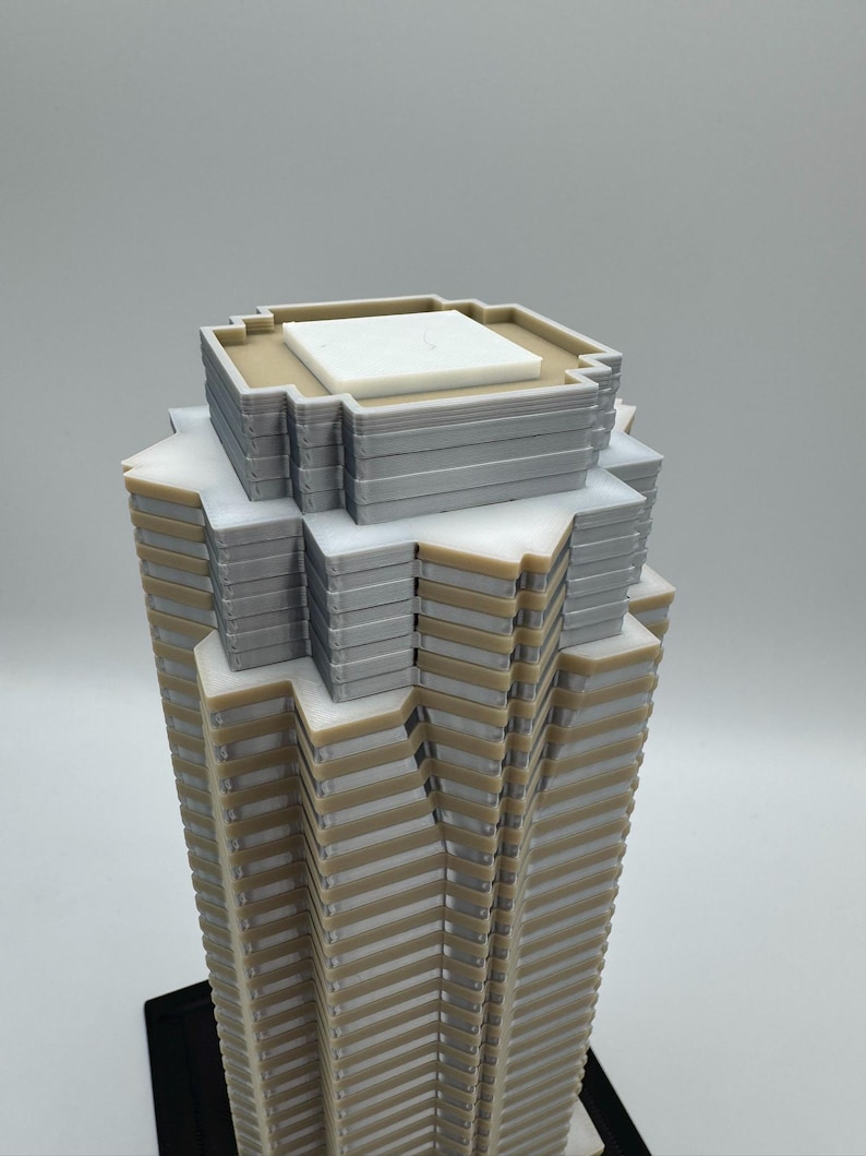 May include: A detailed architectural model of a skyscraper. The model features a layered design with alternating beige and gray sections, topped with a white and gray roof structure. The base is black.