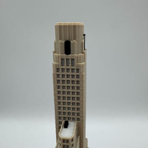 May include: A beige 3D printed architectural model of a skyscraper featuring a stepped design, multiple rows of rectangular windows, and a prominent vertical tower section. The structure includes dark accents on the upper levels and roof.