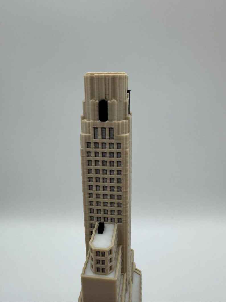 May include: A beige 3D printed architectural model of a skyscraper featuring a stepped design, multiple rows of rectangular windows, and a prominent vertical tower section. The structure includes dark accents on the upper levels and roof.