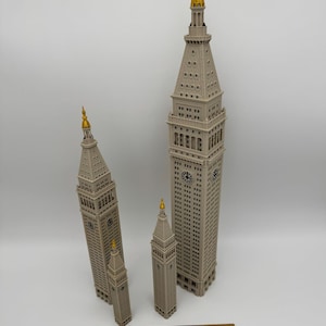 May include: Three beige architectural models of the Metropolitan Life Insurance Company Tower, each featuring a gold-colored spire and clock faces. A wooden ruler is placed at the base, showing the tallest model is approximately 12 inches tall.