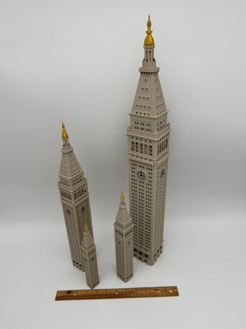 May include: Three beige architectural models of the Metropolitan Life Insurance Company Tower, each featuring a gold-colored spire and clock faces. A wooden ruler is placed at the base, showing the tallest model is approximately 12 inches tall.