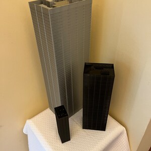May include: Three miniature skyscrapers made of gray and black plastic blocks. The tallest skyscraper is gray and the two smaller skyscrapers are black.